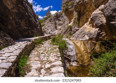 Stone path by a small stream of water between beautiful rocky walls made of limestone. Barranc del Cint trail in the La Sierra de Mariola natural park in Alcoy, Alicante province, Spain, Montcabrer