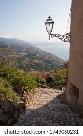 A stone path leading down through a Sicilian village with hills and a misty ocean in the distance