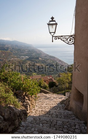 A stone path leading down through a Sicilian village with hills and a misty ocean in the distance