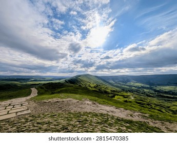 Stone path on Mam Tor ridge overlooking green hills and valley under cloudy blue sky