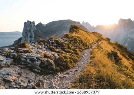 Stone path winds along a grassy alpine ridge toward the setting sun with jagged peaks and glowing haze in the distance.