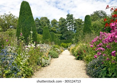 Stone Pathway Leading To A White Bench, With Cottage Colourful Flowers In Bloom On Both Sides, Shaped Conifers, Shrubs And Tall Trees In An English Garden On A Sunny Summer Day