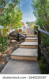 A stone pathway with steps surrounded by greenery and rocks, featuring a wooden railing, thatched umbrella, and lounge chairs under a bright blue sky.