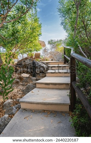 A stone pathway with steps surrounded by greenery and rocks, featuring a wooden railing, thatched umbrella, and lounge chairs under a bright blue sky.