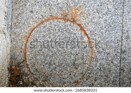 Stone paving displays a circular rust colored mark, a metal ring has left a print, the circle sits at the center of a square tile, rough grains show through, weather and time stain the outdoor surface