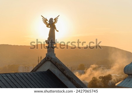 Stone sculpture of an angel with golden wings standing on the rooftop of a baroque temple at sunset.