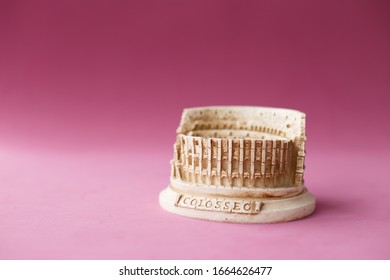 Stone Statuette Of The Coliseum Building In Rome In Italy. Concept Of Dangerous Infection With The Chinese Coronavirus Covid-2019. Pink Background. Beautiful Memory Souvenir For Travelling Tourists.