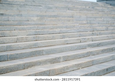 Stone steps are arranged in a repeating pattern, rising up towards a building at the top. The steps are made of gray stone and show some wear from use over time. The weather appears cloudy, adding a neutral tone to the scene. There are no people visible on the steps, and the surrounding area looks clean and well-maintained. This location seems to be part of a public space in a city. Overall, the focus is on the structure and design of the steps.