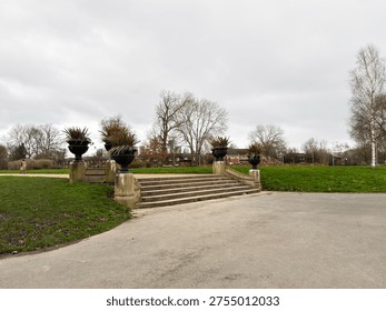Stone steps flanked by large urn planters lead into a quiet Alexandra Park in Manchester on an overcast day.
