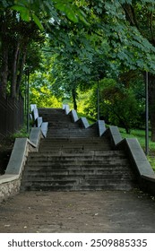 Stone steps with a zigzag railing ascend through a park lined with lush green trees and black lamp posts, creating a serene and natural outdoor environment.