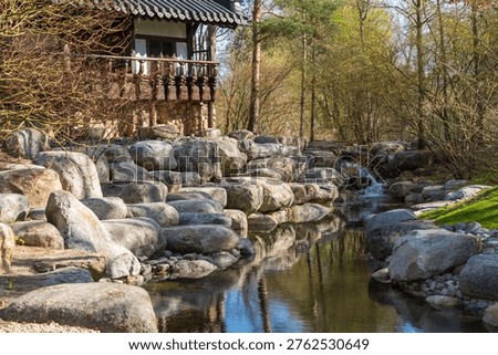 Stone stream flows gently through a tranquil garden. Rocks line the water's edge, forming natural barriers. Traditional wooden structure overlooks the scene