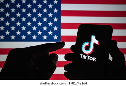 Stone / United Kingdom - July 22 2020: TikTok logo seen on the silhouette of smartphone hold in a hand with blurred American flag on the back. Real photo, not a montage.