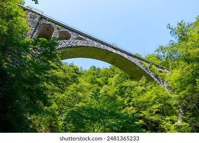 The stone Vintgar Gorge Viaduct in Slovenia showcased in a low-angle view, revealing its historic arch and stone construction, framed by lush green trees in the Julian Alps. - Powered by Shutterstock