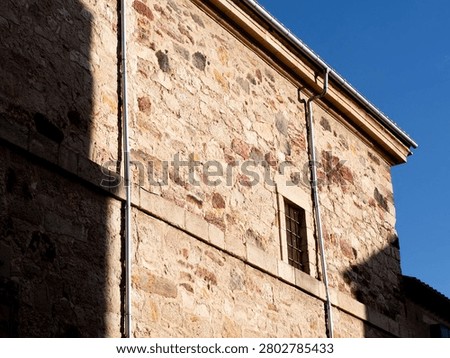 Stone wall facade featuring a window and drainpipe with shadows and clear blue sky