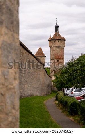 A stone wall and tower stand guard over a path and parked cars under a cloudy sky.