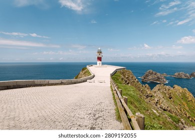 Stone-paved path leads to lighthouse perched on rocky cliff. Ocean stretches below, and sky is clear. Lighthouse on a cliff face on a sunny day. Concept of nature and beauty.