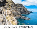 Stone-paved trail with rustic wooden posts and rope railings hugs a rugged volcanic cliff coastline at Poris de la Candelaria, La Palma, Canary Islands, Spain