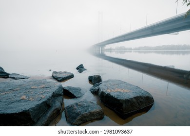 Stones in the water in the foreground and cable-stayed bridge over foggy morning river Dnipro. Kyiv. Ukraine. Riverbank low-angle shot at Kyiv.