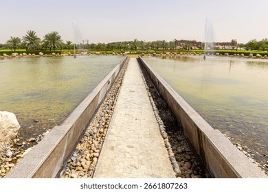 Stony bridge walking path through the pond with fountains in Quranic Park, Dubai, United Arab Emirates, lush green gardens with date palm trees in the background.