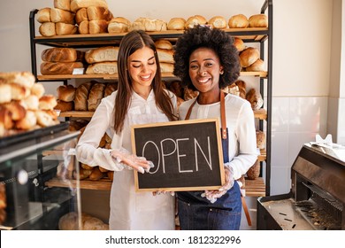 Stop by for homemade delicious bread! Loving young women embracing holding a sign together on the opening day of their small business - couple owners of the bakery smiling to the camera