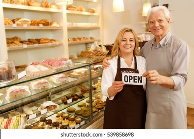 Stop by for homemade delicious bread! Loving senior couple embracing holding a sign together on the opening day of their small business senior couple owners of the bakery smiling to the camera - Powered by Shutterstock