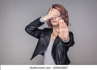 Stop, I dont want to see. Portrait of blocking girl with short hairstyle and makeup in casual style black leather jacket standing with stop gesture. indoor studio shot, isolated on grey background.