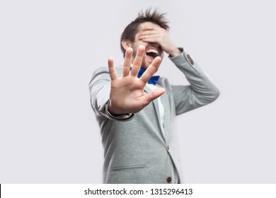 Stop, i don't want to see this. Portrait of shocked or scared young bearded man in casual grey suit standing closed eyes and blocking with hand. indoor studio shot, isolated on light grey background.