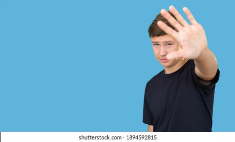 Stop Gesture. Refusal Sign. Offer Rejection. No Thanks. Portrait Of Annoyed Teen Guy In Black T-shirt Showing Hand Open Palm Looking At Camera Isolated On Blue Copy Space Commercial Background.