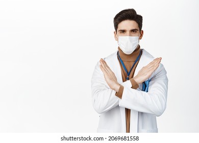 Stop. Healthcare Worker In Medical Face Mask And White Robe, Showing Cross No Gesture, Disapprove Action, Prohibit Smth, Standing Over Studio Background