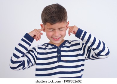 Stop Making This Annoying Sound! Unhappy Stressed Out Little Cute Caucasian Boy Kid Wearing Stripped T-shirt Against White Wall Making Worry Face, Plugging Ears With Fingers, Irritated With Loud Noise