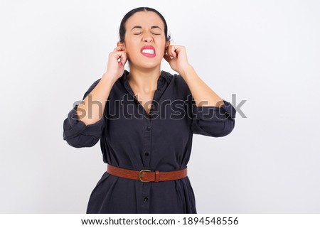Stop making this annoying sound! Unhappy stressed out young beautiful Arab woman wearing gray dress against white studio background making worry face, plugging ears with fingers.