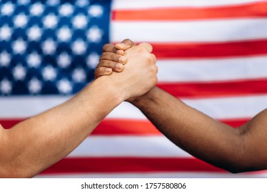 Stop Racial Discrimination! Black African American Male Hand And White Caucasian Male Hand Make A Handshake In Agreement Of Friendship Against The Background Of The Flag Of USA