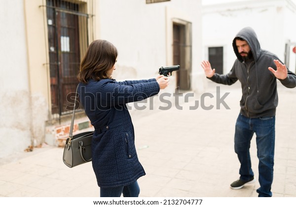 Stop right there! Angry young woman pointing a gun\
to a criminal thief. Young man and mugger putting his hands up on\
the street