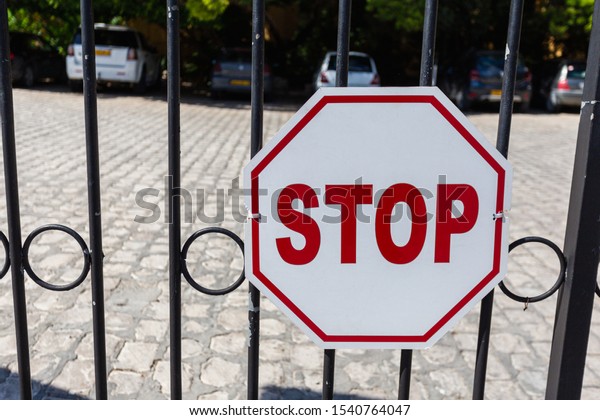 Stop sign on the fence in front of the Parking\
lot. Close up of a red hexagonal stop sign on a metal fence with\
parked cars in the\
background