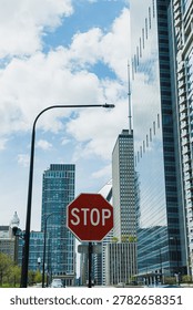 A stop sign is placed at an intersection surrounded by tall buildings and clear blue skies. The scene shows a busy urban environment and displays a typical city layout.