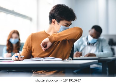 Stop Spreading Virus Concept. Asian male student couching in his elbow, sneezing in sleeve during lecture in university. Guy wearing protective medical mask, sitting at desk in classrom - Powered by Shutterstock