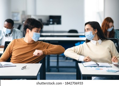 Stop Spreading Virus Concept. Smiling Diverse Students Wearing Protective Face Masks Greeting Each Other And Bumping Elbows At Classroom. Man And Woman Sitting At Desks, Studying In High School