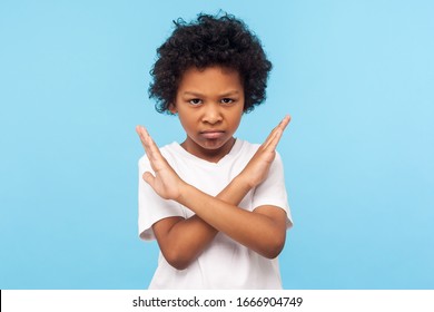 Stop, I'm warning. Portrait of angry determined little boy with curls crossing hands and looking at camera with aggression, showing stop gesture, way prohibited. indoor studio shot blue background