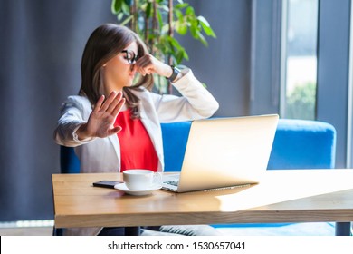 Stop, You Stink! Portrait Of Beautiful Stylish Brunette Young Woman In Glasses Sitting, Pinching Her Nose And Showing Stop Gesture Because Bad Smell. Indoor Studio Shot, Cafe, Office Background.