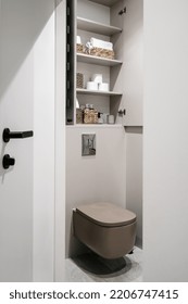 Storage Closet With Paper Rolls, Clean Towels, Hygienic Accessory In Bathroom Over Built In Toilet Bowl Installed On Wall. Opened Doors On Cupboard With Household Items. Restroom Interior