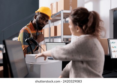 Storage room supervisor checking customer order on computer, discussing shipping details with african american employee in warehouse. Diverse team working at goods quality control in storehouse