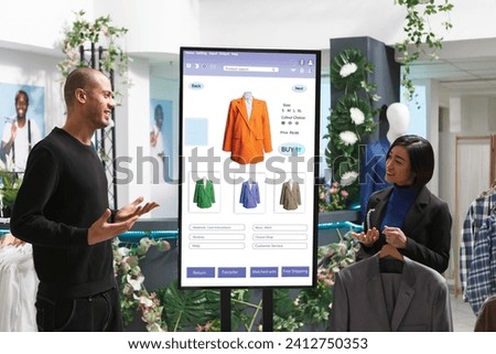 Store associate displays items to man in front of digital kiosk, selecting merchandise from clothing boutique. Caucasian male customer converses with female employee about products.