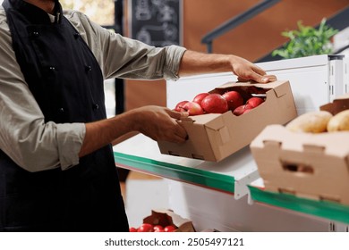 Store worker wearing black apron handles small crate of several red apples on a shelf in grocery store. Close-up of male vendor placing box of fresh fruits on white racks of eco friendly supermarket. - Powered by Shutterstock