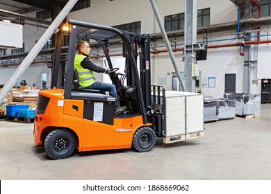 Storehouse Employee In Uniform Working On Forklift In Modern Automatic Warehouse. Boxes Are On The Shelves Of The Warehouse. Warehousing, Machinery Concept. Logistics In Stock.