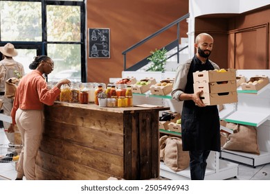 Storekeeper carrying delivered crates full of freshly harvested produce for inventory in eco friendly store. Young male vendor grasping boxes of organic products while customer browses the shop. - Powered by Shutterstock