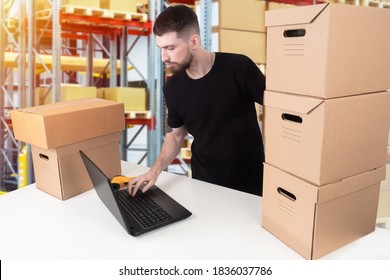 Storekeeper in the warehouse. Warehouse account. This man gives orders from the warehouse. The storekeeper works on a laptop. A man next to cardboard boxes and a laptop. - Shutterstock ID 1836037786