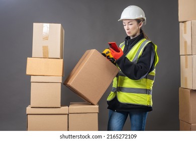 Storekeeper Woman. Warehouse Manager With Phone And Laser Scanner. Storekeeper Is Holding Mobile Phone. Employee Of Warehouse Company Scans Codes On Box. Girl In Yellow Vest Among Cardboard Boxes