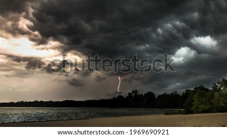 Storm clouds and lightning strike over Lake Ronkonkoma, New York on a rainy day.  Located on Long Island.  The beach can be seen where the sand meets the water at the shoreline. There is some sunlight