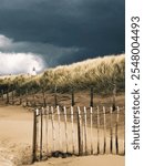 Storm clouds loom over sandy dunes topped with wild grass, framing a rustic wooden fence and a distant lighthouse. A serene yet dramatic coastal landscape showcasing the raw beauty of nature.