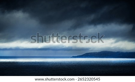 Storm clouds over the Côte d’Azur with sunlight breaking through to illuminate the sea. Dramatic seascape with moody skies and a distant coastal silhouette.